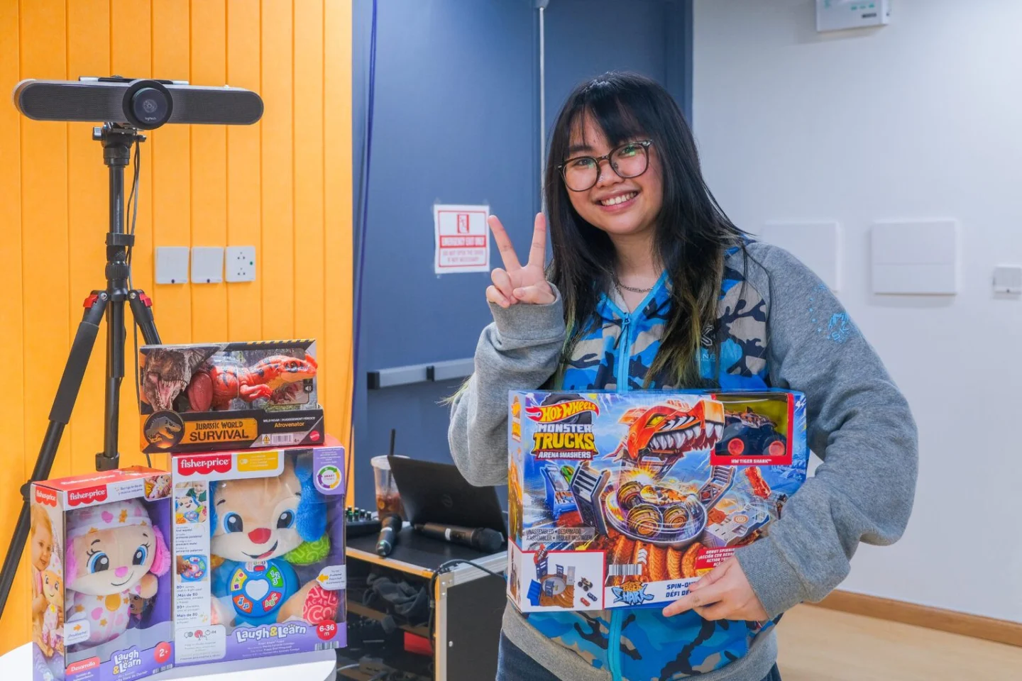 A smiling BUV student holds a Hot Wheels toy set and makes a peace sign beside donated toys and a camera on a tripod.