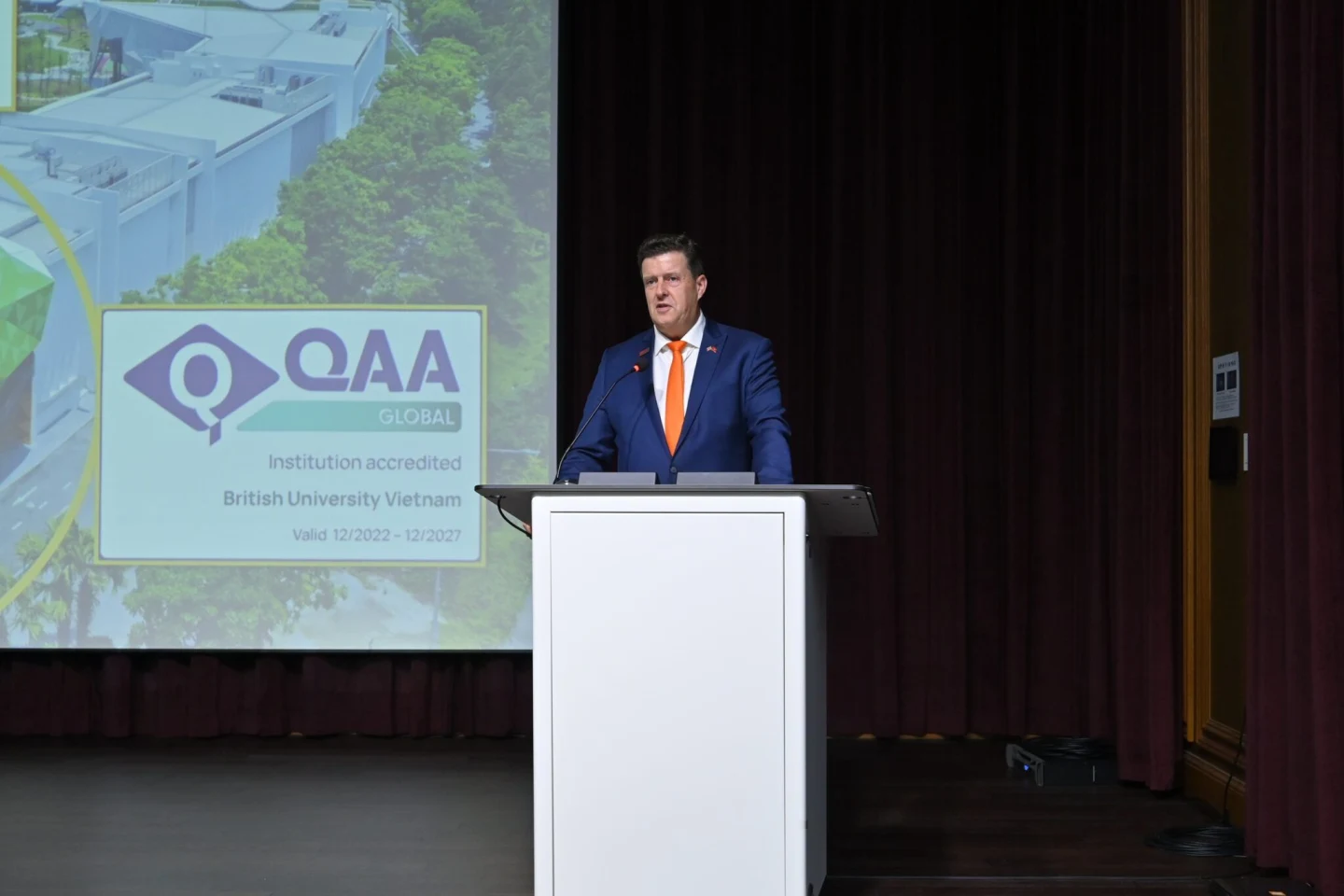 A speaker in a navy suit and orange tie stands at a podium beside a presentation screen displaying the QAA Global Quality Licence accreditation.