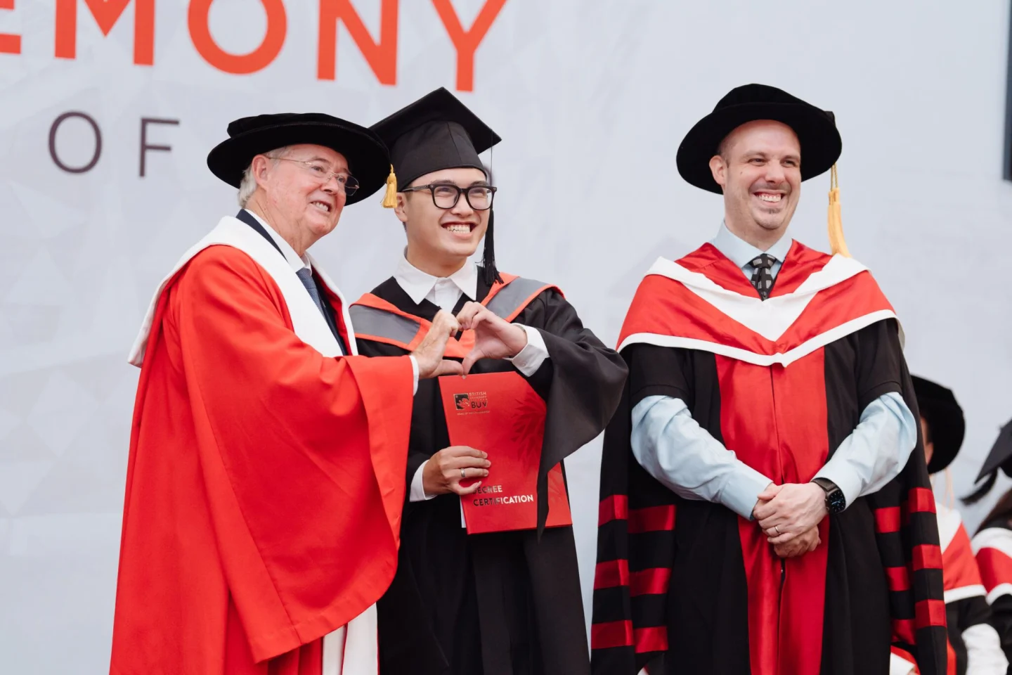 A graduate holding a red diploma makes a heart gesture with a university official, while another academic in ceremonial robes smiles beside them.