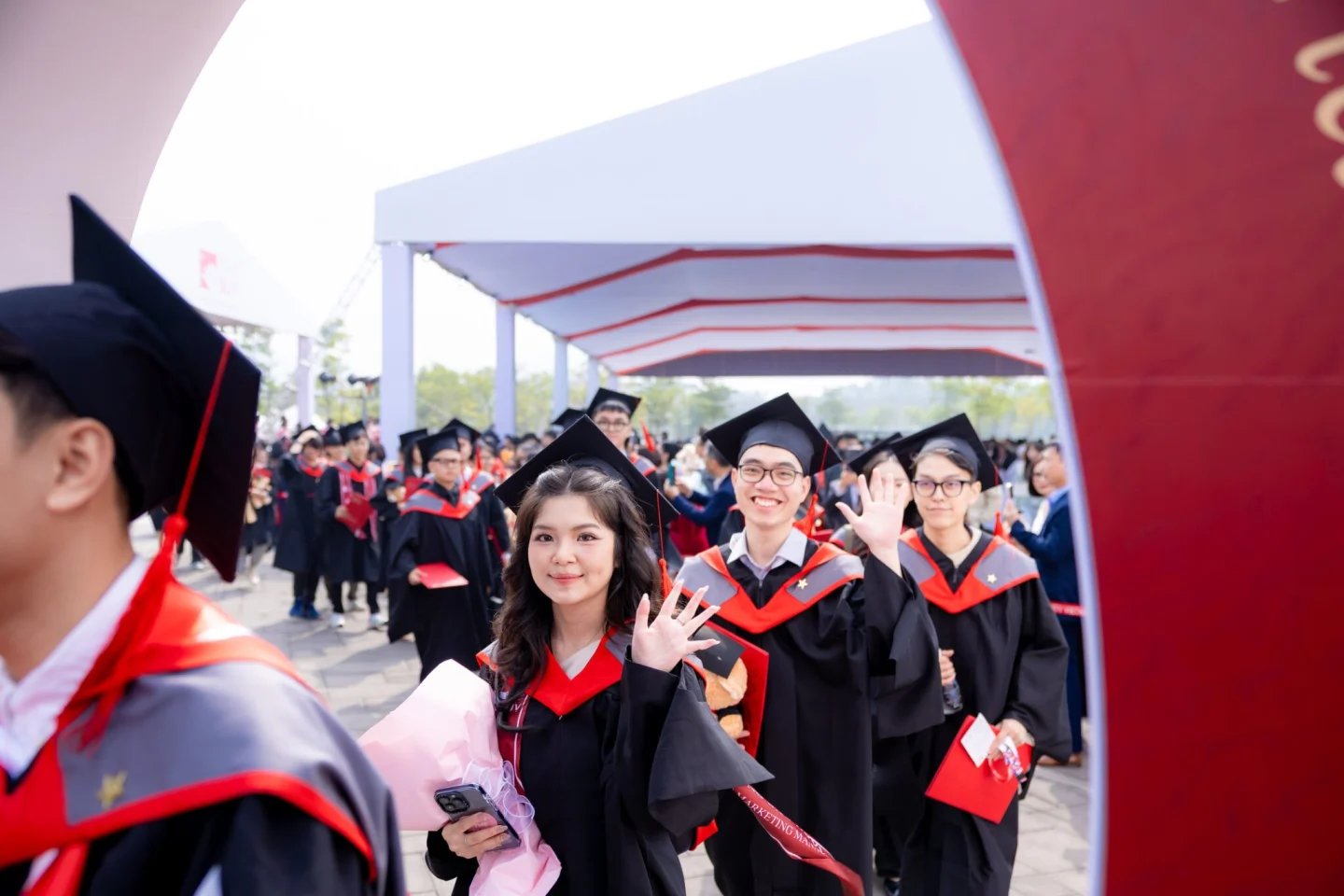 BUV graduates in black gowns with red trim smile and wave while walking in a procession at an outdoor graduation ceremony.