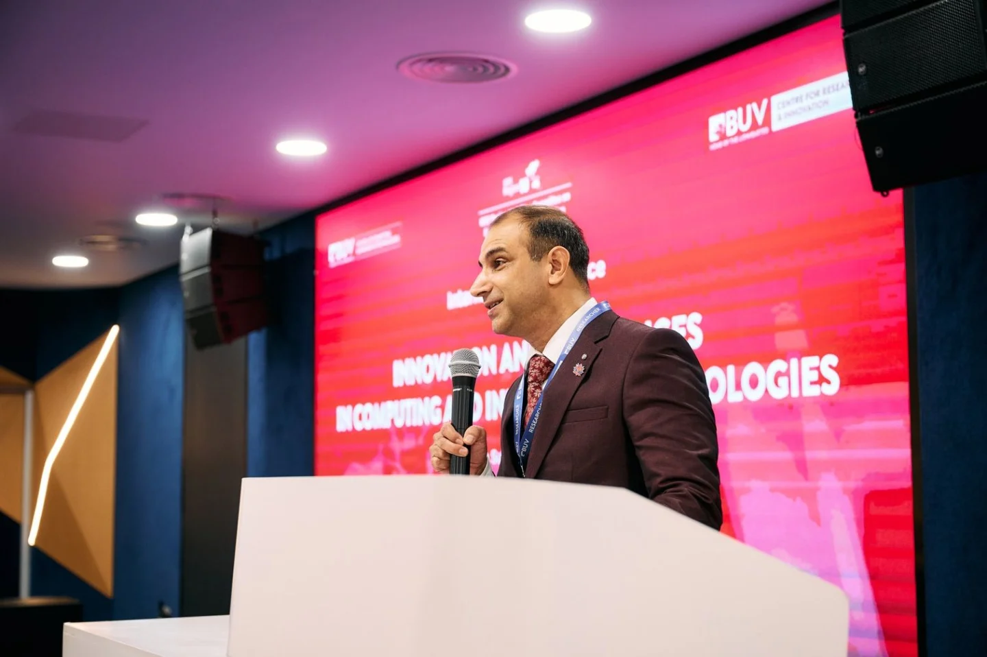 A suited speaker addresses the audience from a white podium at a BUV event, with a red conference backdrop behind him.