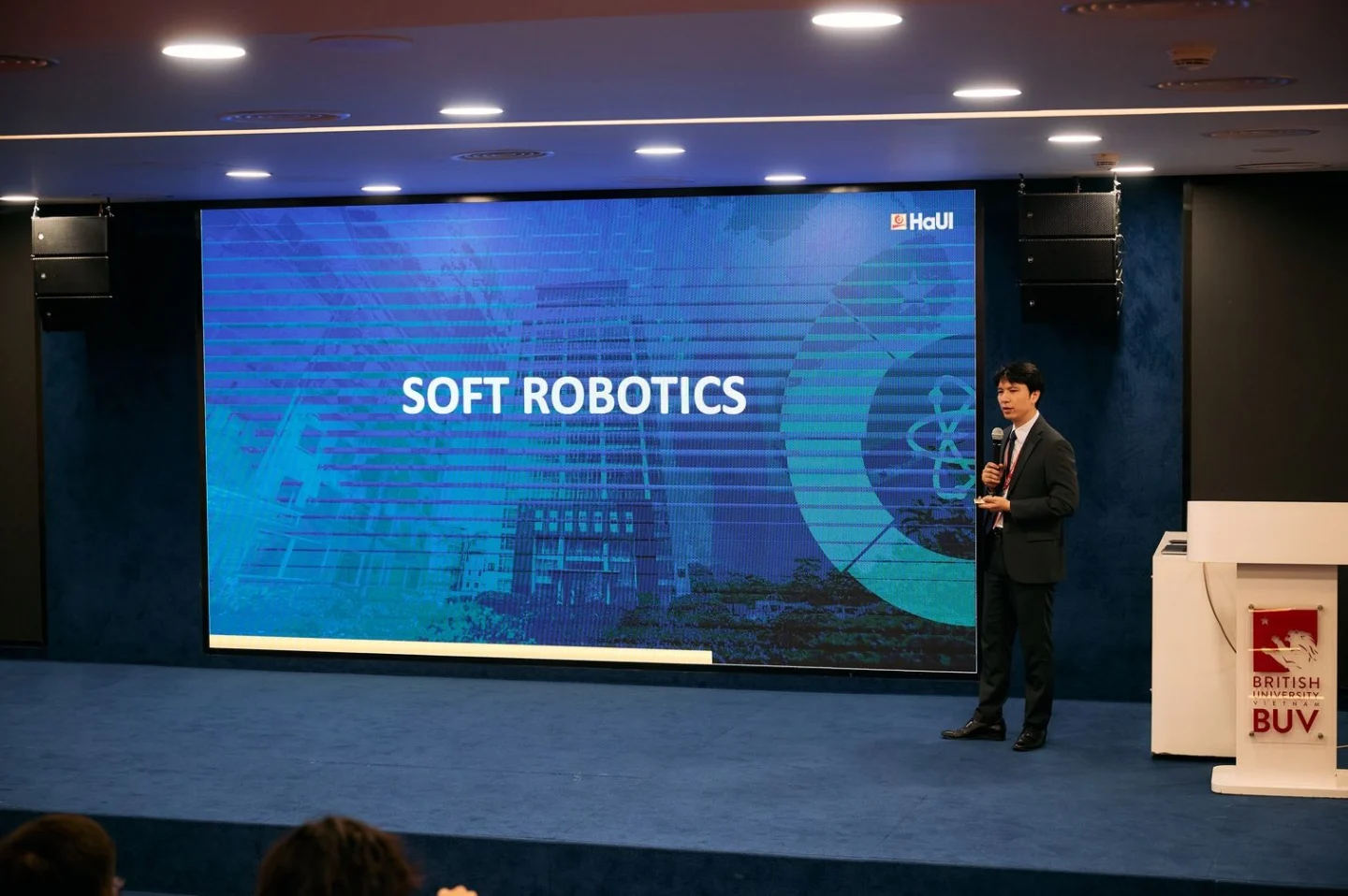 A BUV presenter speaks on stage beside a large screen displaying “SOFT ROBOTICS” during a lecture or academic event.