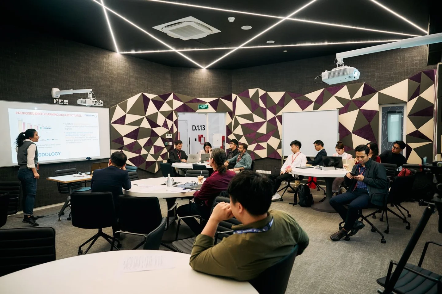 A lecturer presents to students seated around tables in a modern BUV classroom with geometric wall patterns and ceiling lights.