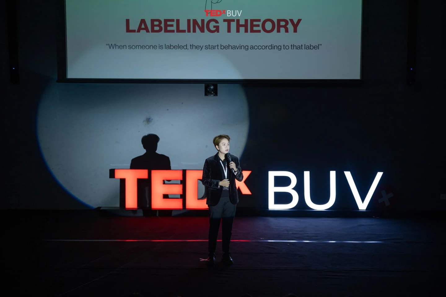 A speaker presents on stage at TEDxBUV beneath a slide titled “Labelling Theory.”
