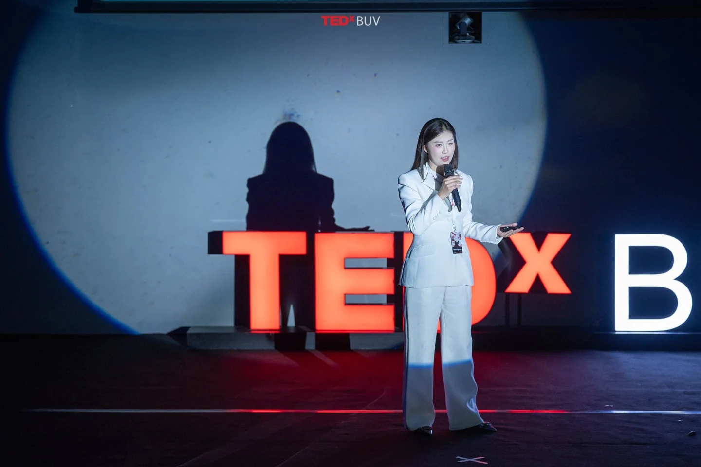 A female speaker in a light grey suit delivers a TEDx talk on stage at British University Vietnam.