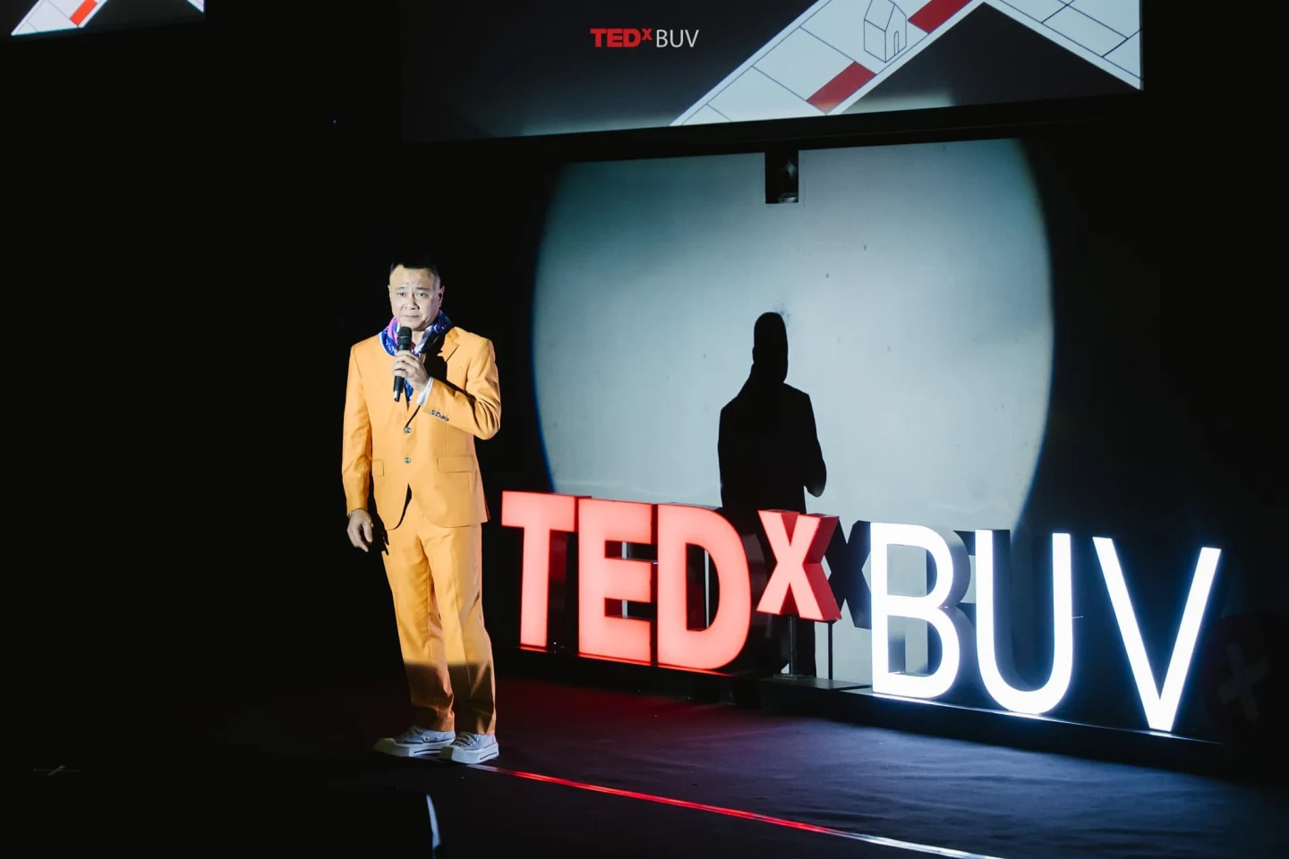 A speaker in a bright yellow suit delivers a TEDxBUV talk onstage beside large illuminated TEDxBUV letters.