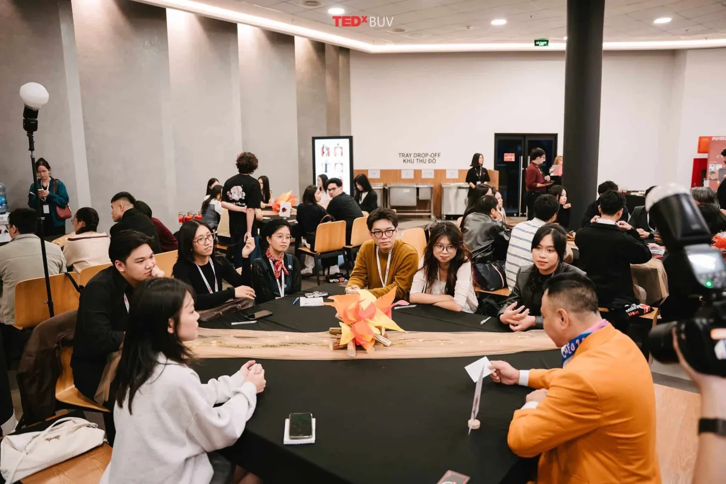Attendees sit around tables during a TEDxBUV networking activity, with an orange origami centerpiece in the foreground.