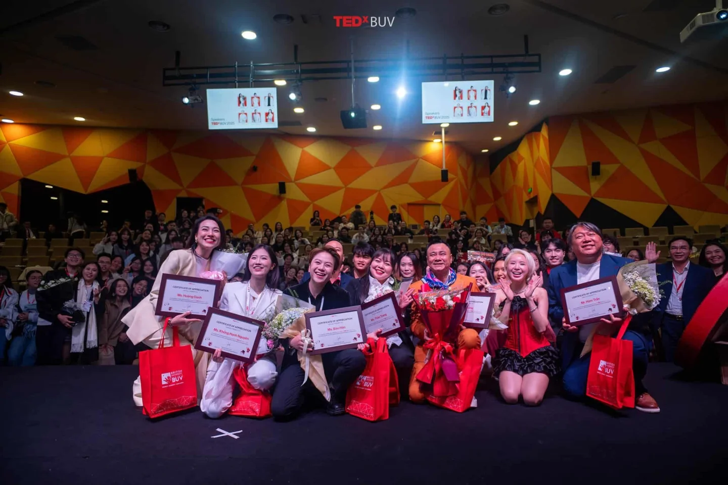TEDxBUV speakers and organizers pose on stage with certificates, flowers, and gift bags in front of a cheering audience.