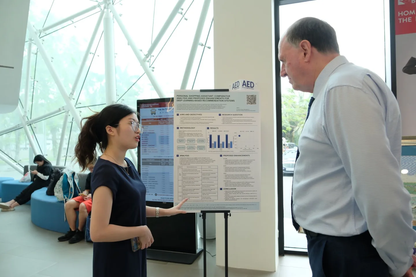 A student presents an AED research poster with charts to a man in a bright, modern university atrium.
