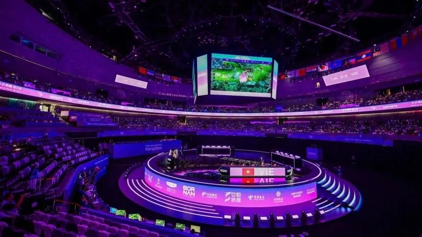 A packed esports arena illuminated in purple, featuring a circular competition stage and large overhead screens displaying live gameplay.