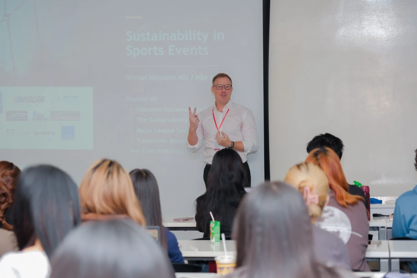 A lecturer presents in front of a projected slide while students listen in a classroom.