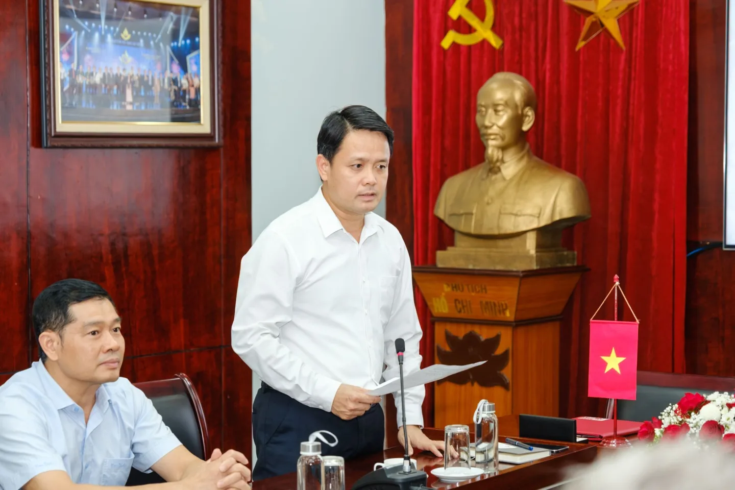 Cán bộ phát biểu tại cuộc họp trong phòng họp Việt Nam, trước tượng Chủ tịch Hồ Chí Minh và quốc kỳ.