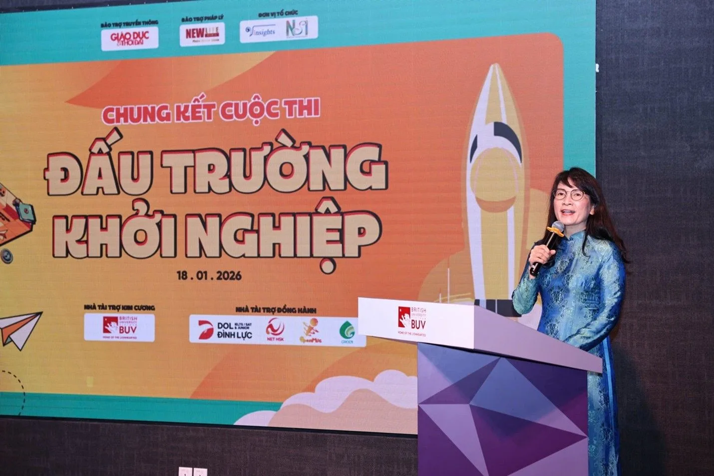 A woman speaks into a microphone at a podium in front of a colorful screen reading “Chung kết cuộc thi Đấu trường khởi nghiệp” (Startup Arena competition final).