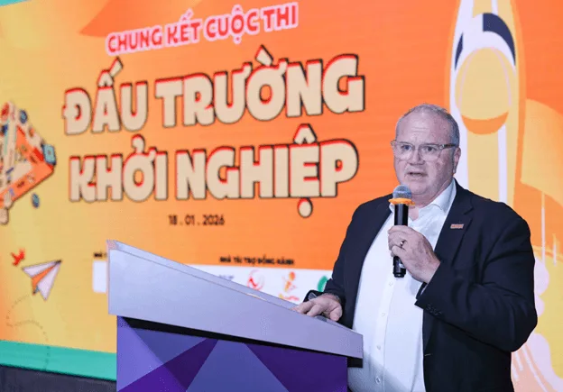 A man in a suit speaks into a microphone at a podium in front of an orange backdrop displaying the Vietnamese text “Chung kết cuộc thi Đấu trường khởi nghiệp” and the date 19.01.2024.