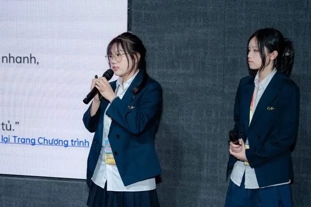 Two female students in navy school uniforms stand in front of a presentation screen; one speaks into a microphone while the other listens.