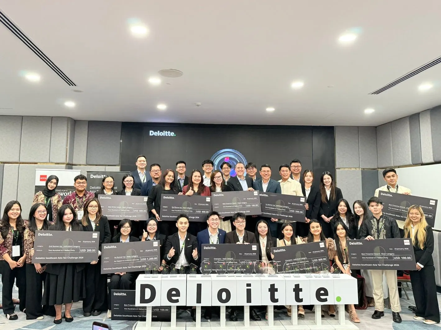 A large group of students and staff pose at a Deloitte office, holding oversized award certificates behind a prominent Deloitte sign.