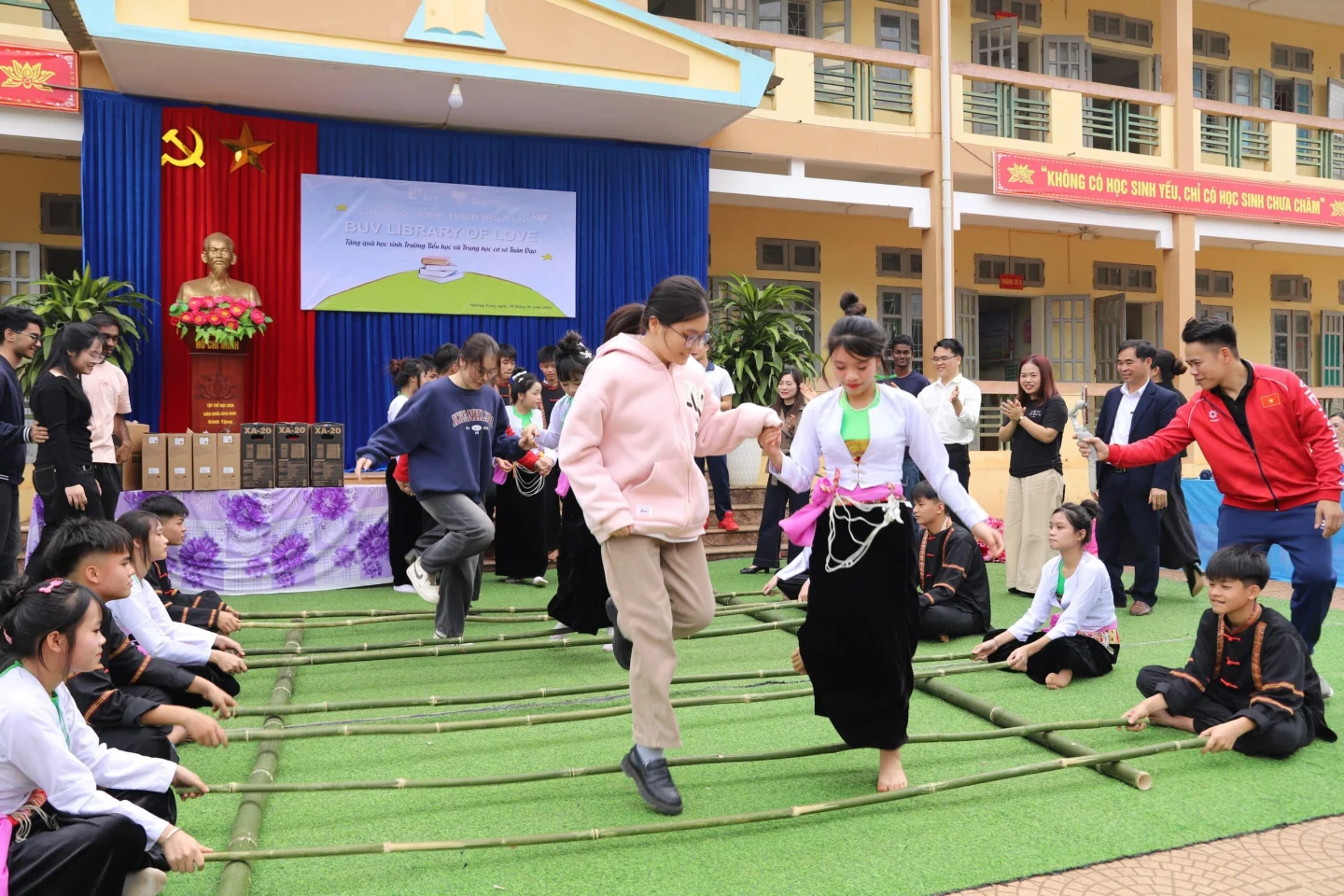 Students participate in a traditional Vietnamese bamboo dance on the steps of a school courtyard, surrounded by seated classmates and an audience.