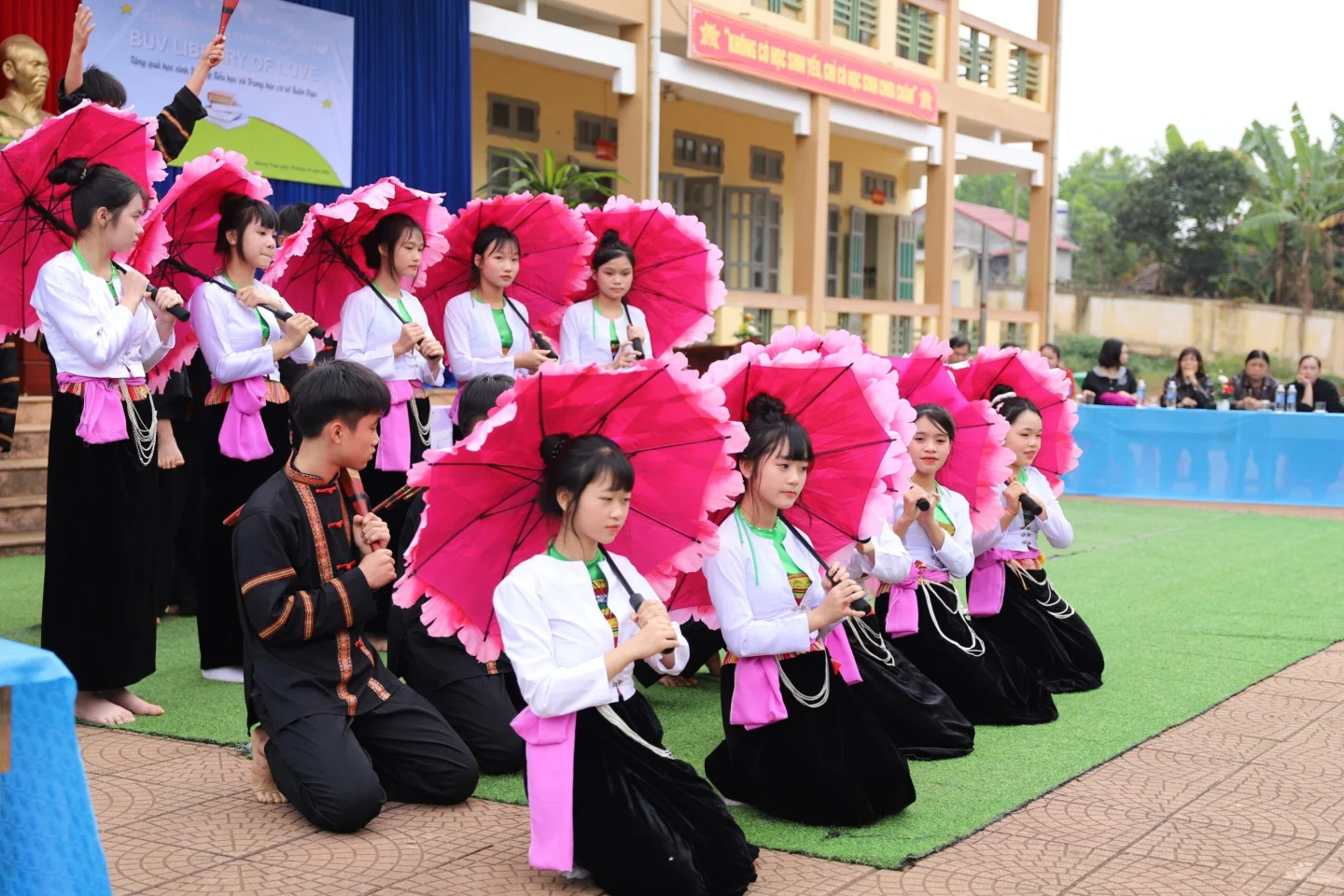 A group of women in traditional Vietnamese attire perform with bright pink parasols in a school courtyard, with several kneeling in the foreground and others standing behind them.