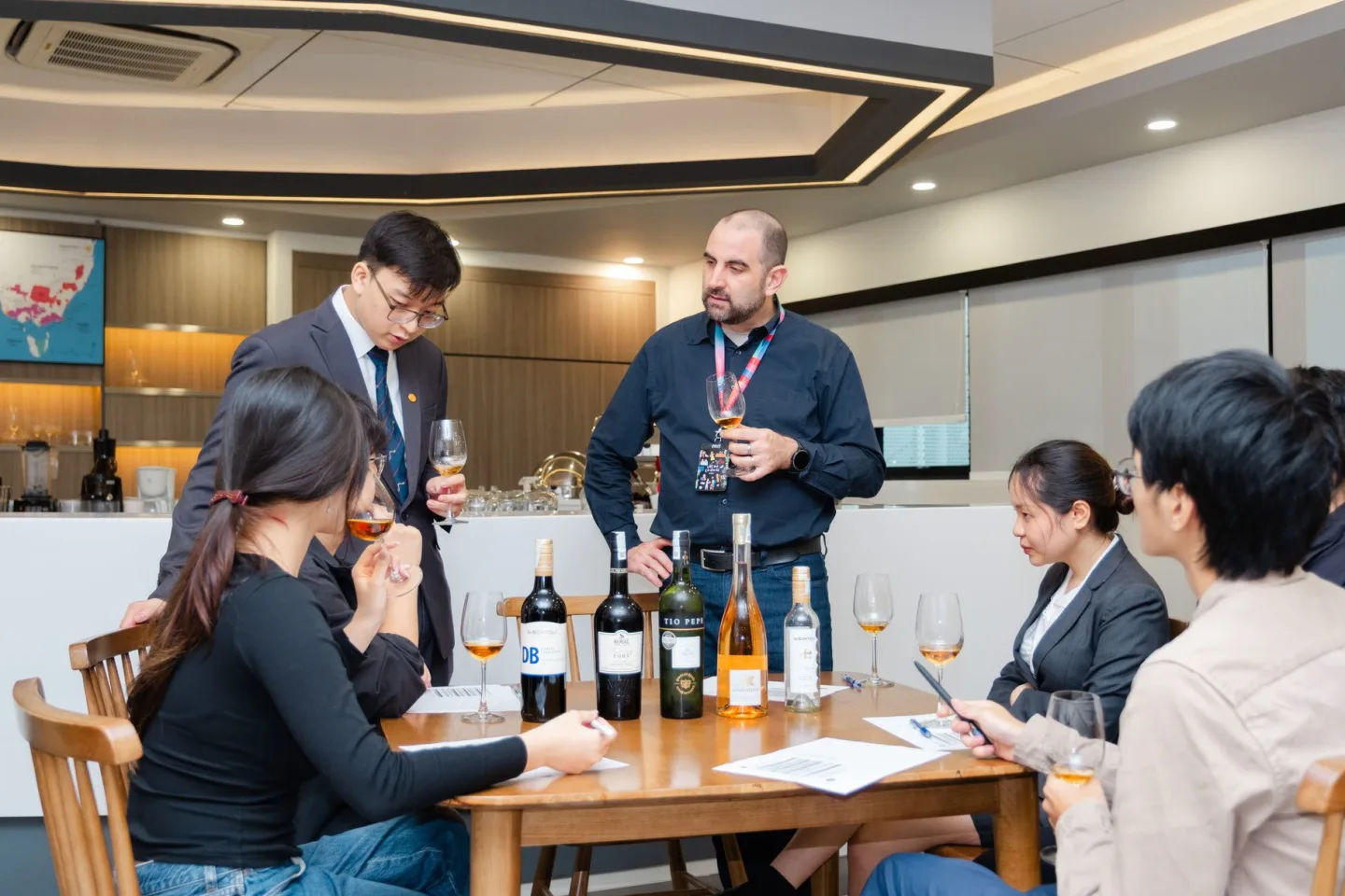 A group of people participates in a wine-tasting session around a table with wine bottles and glasses in a modern room.