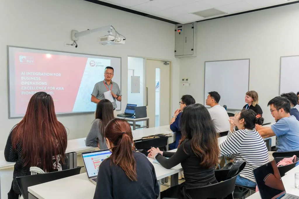 A lecturer presents at the front of a modern BUV classroom while students listen and take notes on laptops.