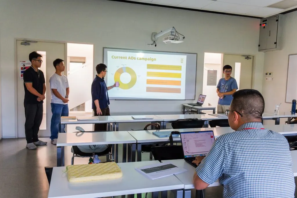 Four students present a “Current Job Campaign” slide with charts to classmates in a modern BUV classroom.