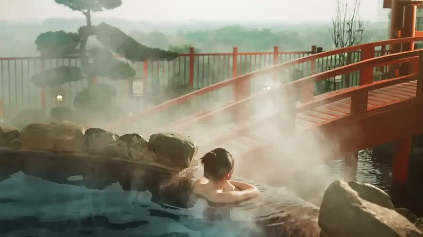 Guest relaxing in the steaming outdoor mineral bath at Mori Onsen, overlooking a red wooden bridge and landscaped surroundings.