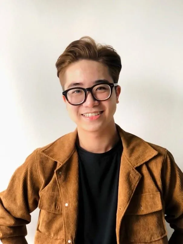 Portrait of a smiling young man wearing black-framed glasses, a black shirt, and a brown corduroy jacket against a plain light background.