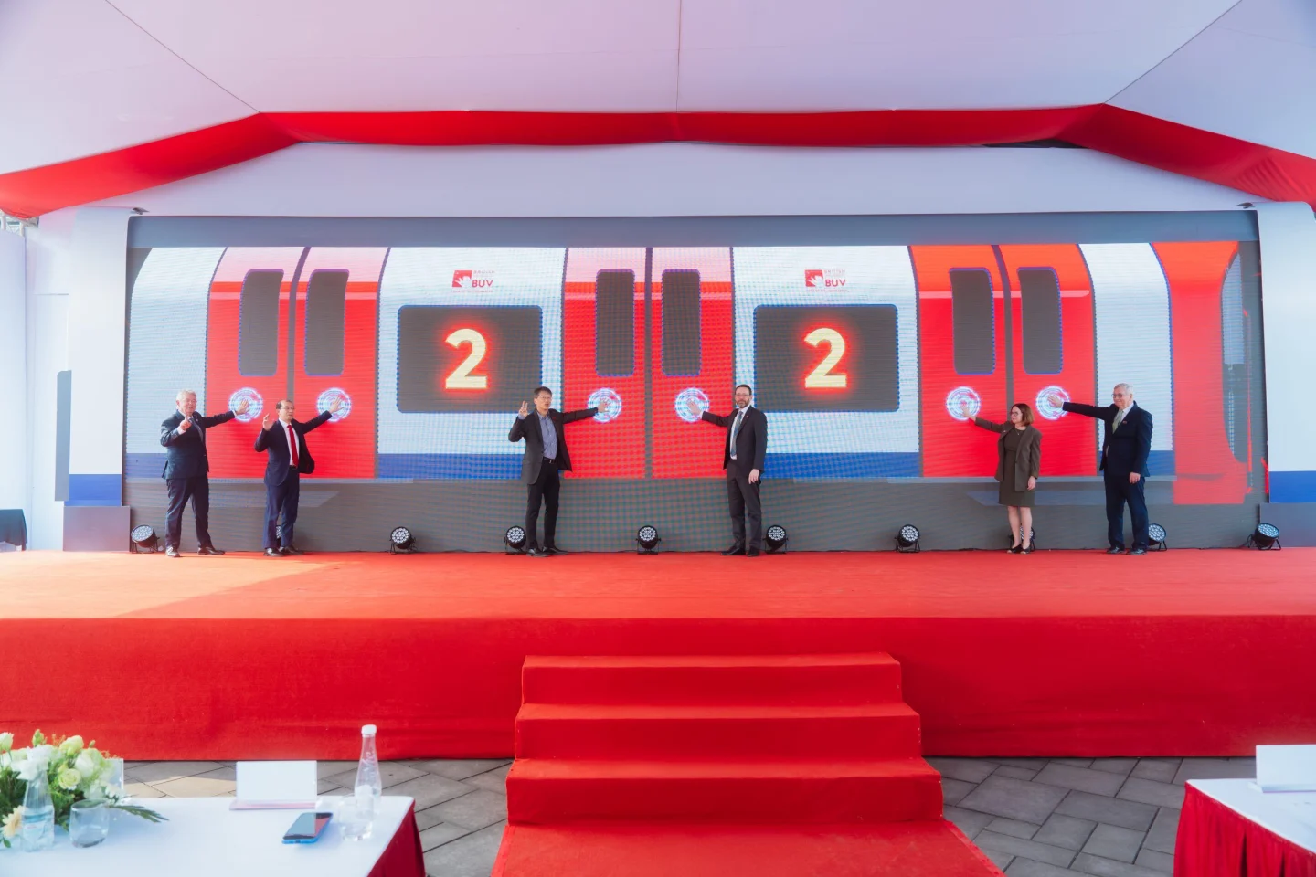 Six officials stand on a red-carpeted stage during a launch ceremony, in front of a large screen depicting a red-and-silver train with illuminated number 2 signs.