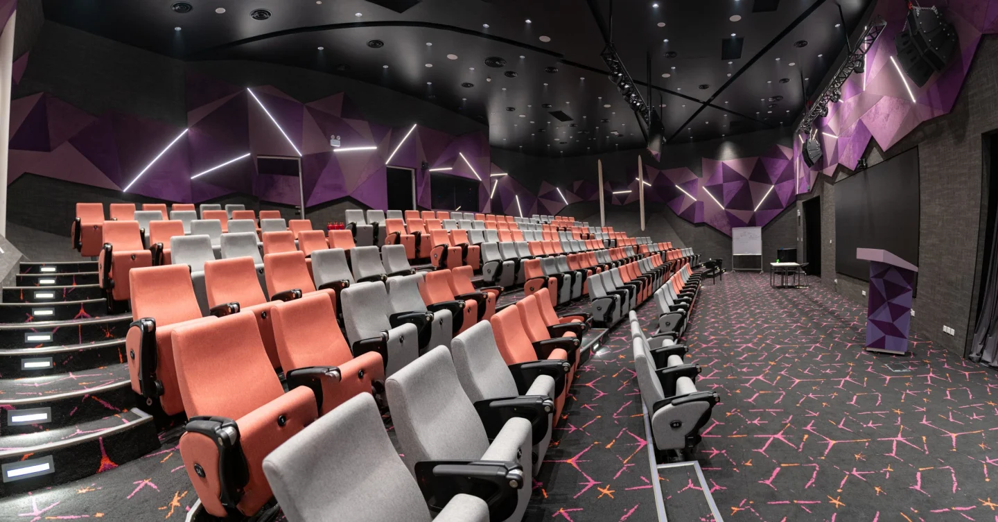 Modern university auditorium with tiered rows of orange and grey seats, geometric purple wall panels, and overhead lighting.