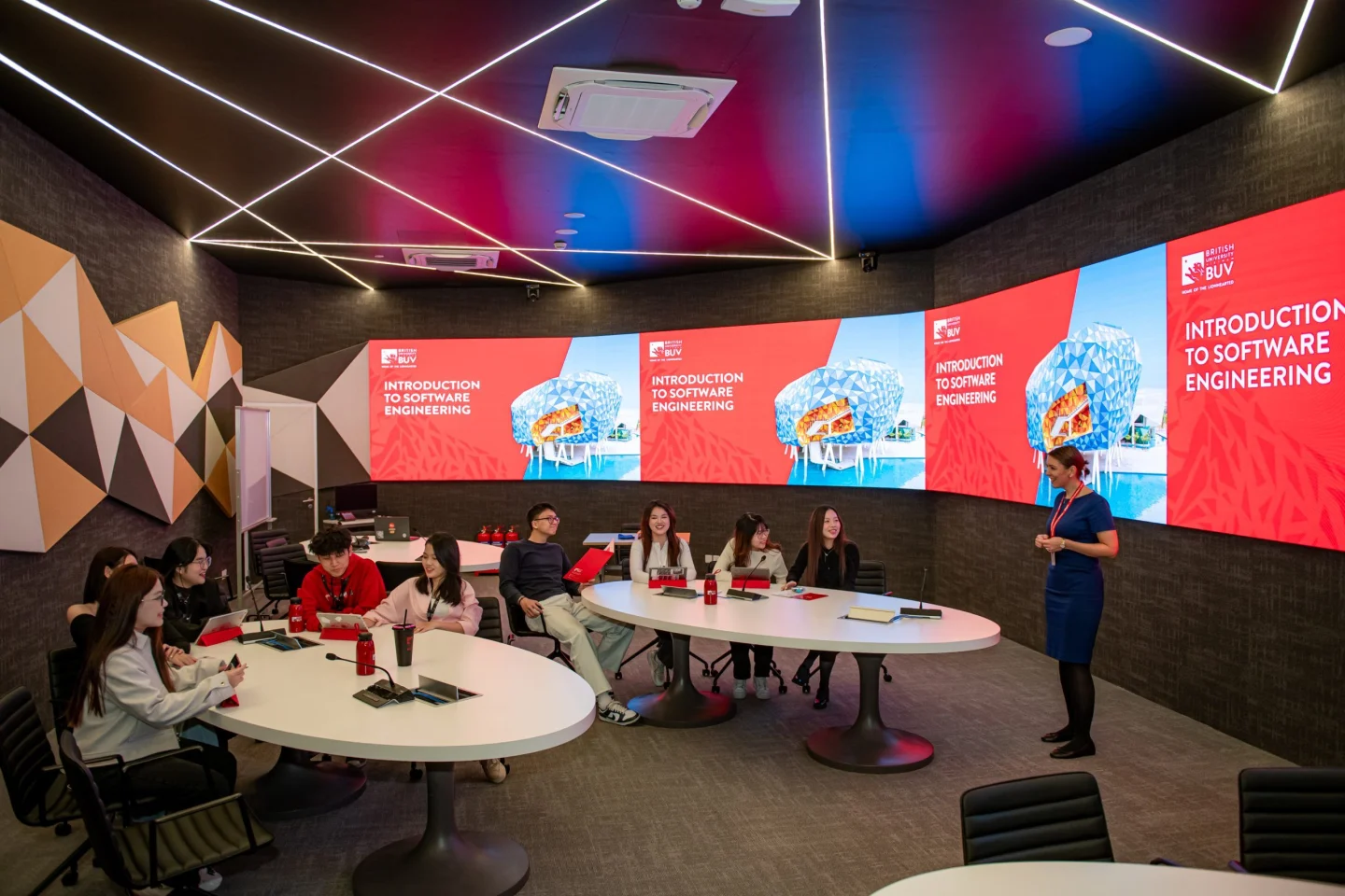 A lecturer presents an Introduction to Software Engineering session to students seated around tables in a modern BUV classroom with large digital screens.