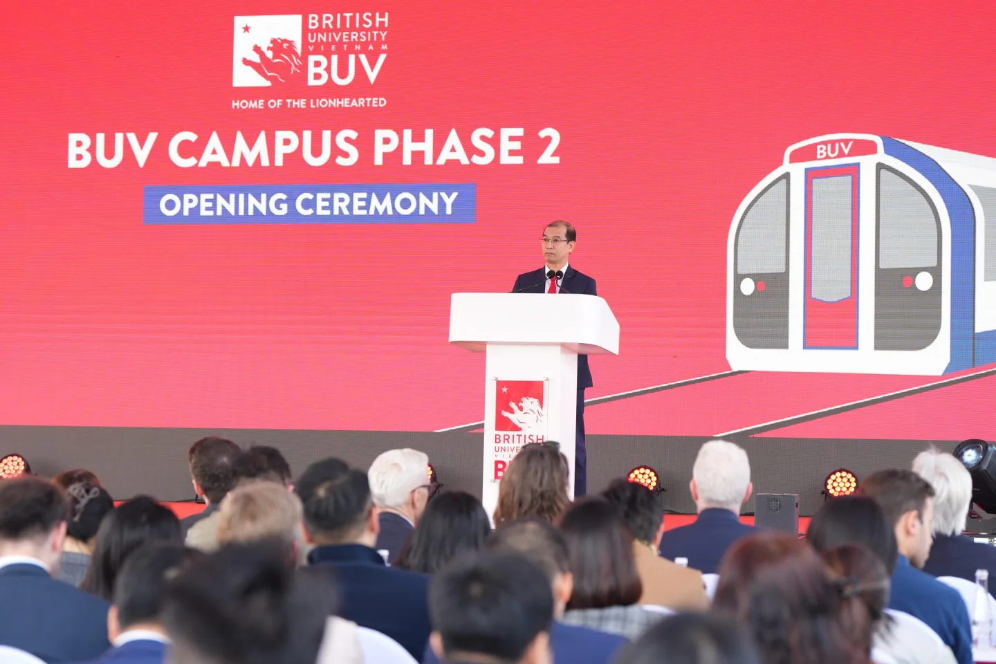 A speaker addresses guests at the BUV Campus Phase 2 opening ceremony, standing at a podium before a red backdrop featuring the British University Vietnam logo and a train illustration.
