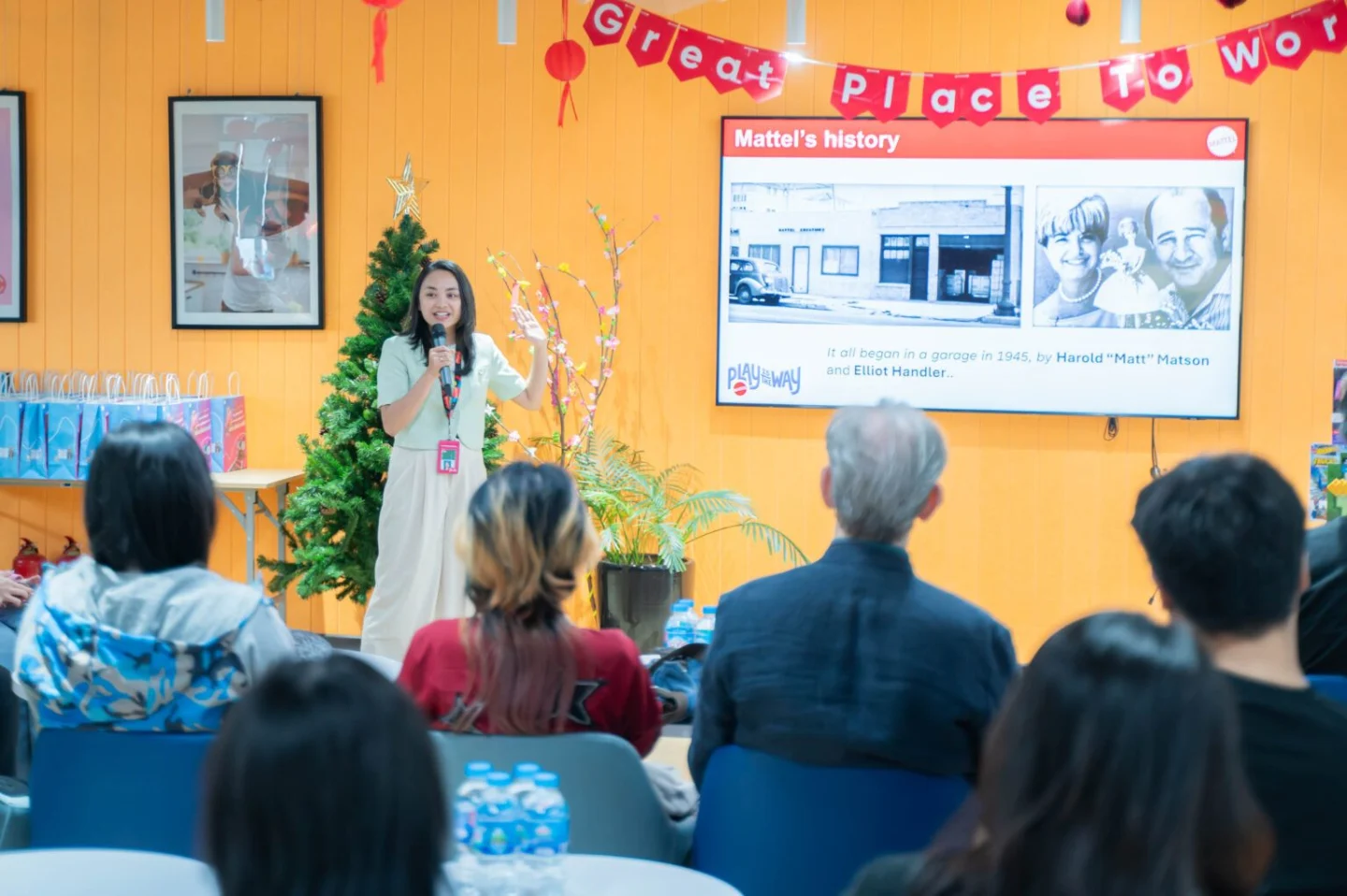 A woman presents Mattel’s history to a seated audience in a colorful room decorated with a “Great Place to Work” banner.