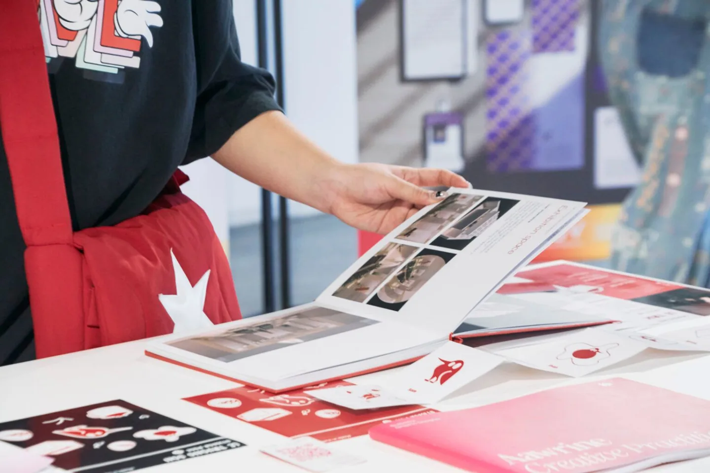A person in a black shirt and red apron reviews printed design materials spread across a table.