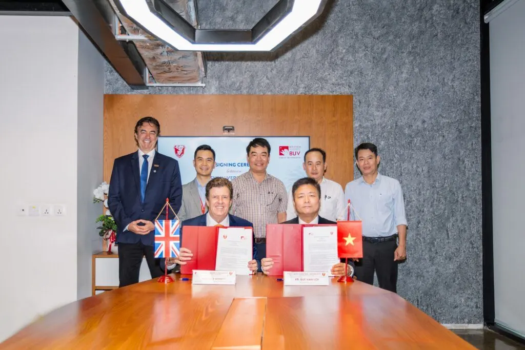 A group photo of several university representatives and officials seated and standing around a table during a formal document signing ceremony with flags of the UK and Vietnam.