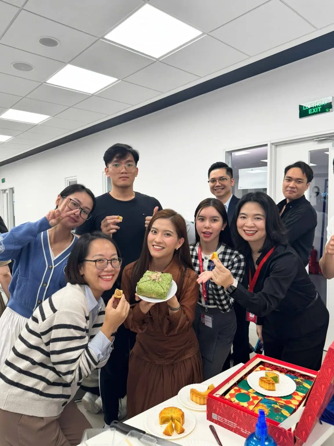 A group of staff and students pose indoors around a decorated red cake box, with one woman holding a plate of green-frosted cake.