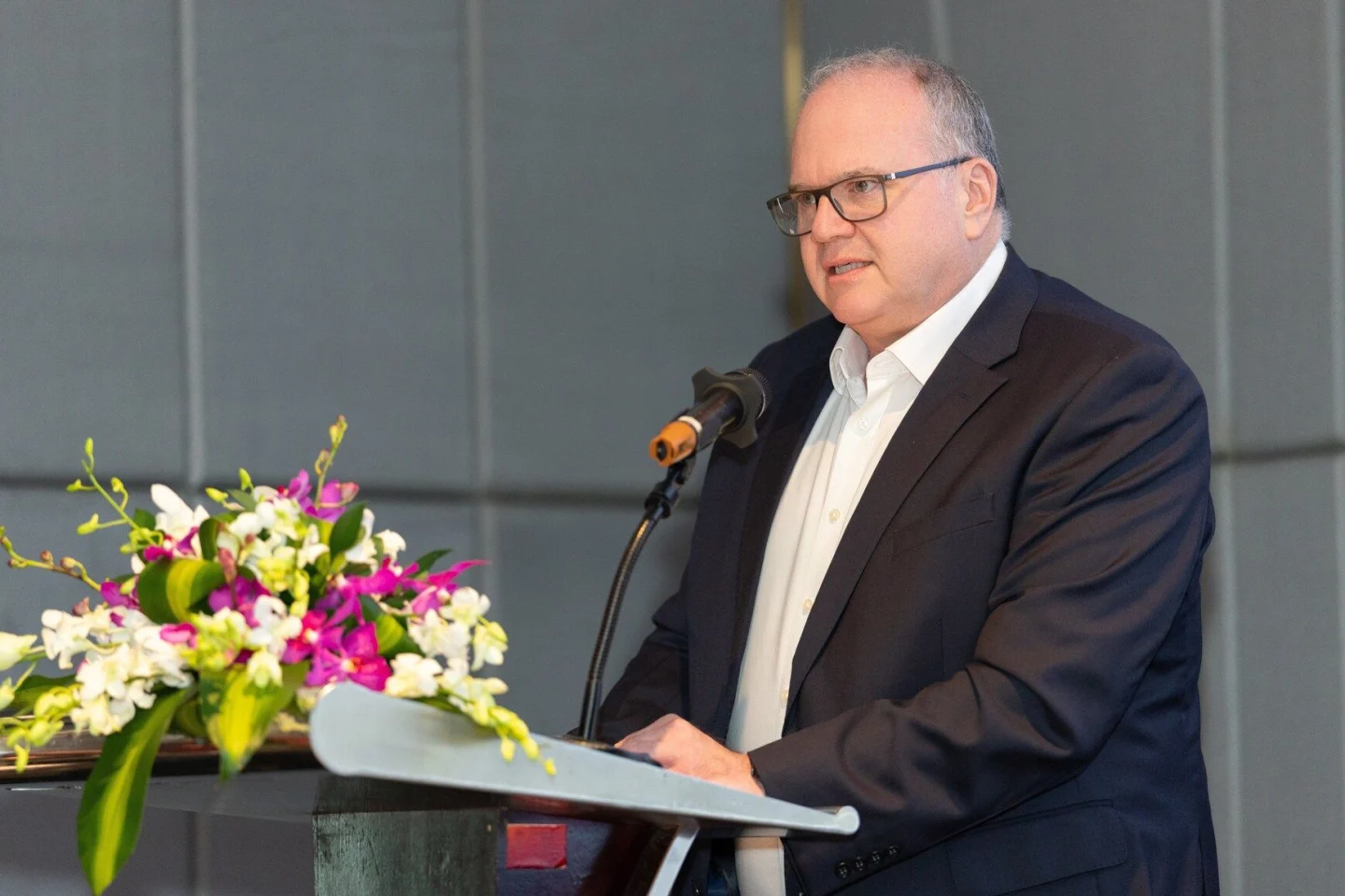 A speaker in a dark suit addresses an audience from a lectern beside a floral arrangement at a BUV event.