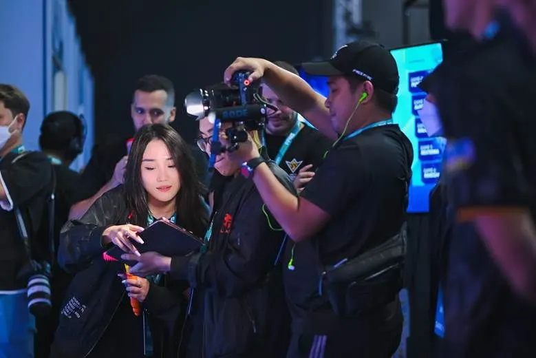 A woman in graduation attire stands as a camera crew records her at an event, with photographers and equipment visible in a blue-lit setting.