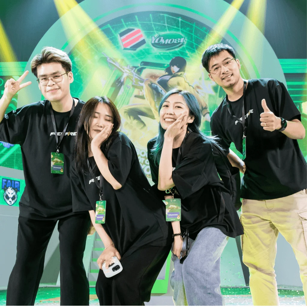 A group photo of four young people in black shirts posing and smiling at an event, with a colorful event banner in the background.