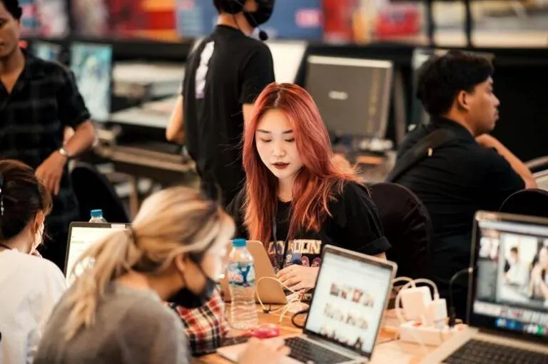 Alt text: A young woman with dyed orange-red hair studies and works at a computer in a classroom or workshop setting.