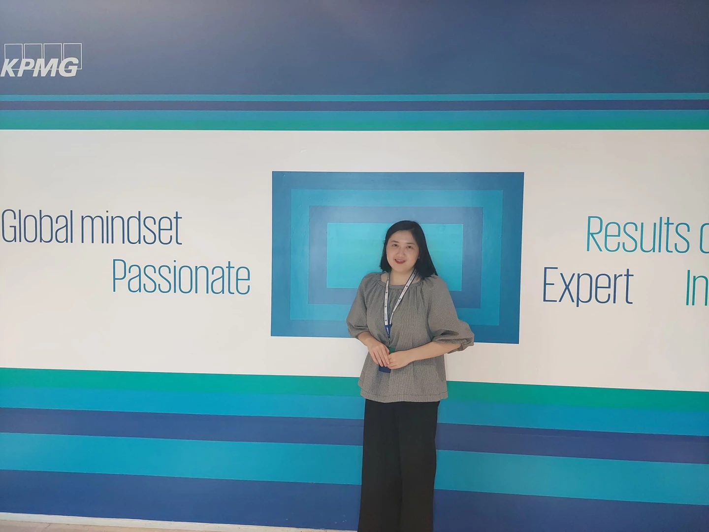 A woman stands at a podium giving a presentation on a conference stage with a screen showing “Global mindset, Passionate” and “Results” in the background.