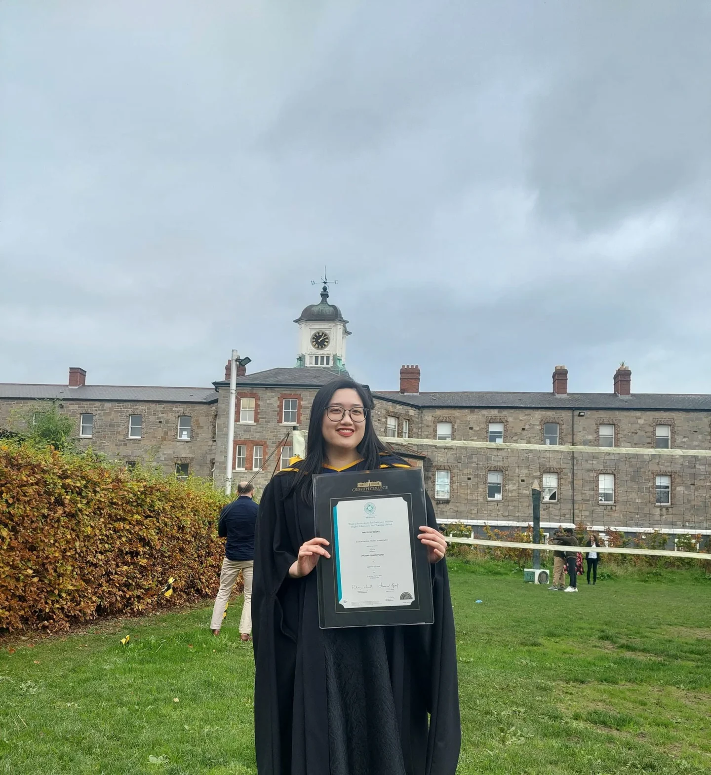 A female graduate in a black graduation gown and cap holds a framed diploma outdoors on a university campus, with historic buildings and a clock tower in the background.