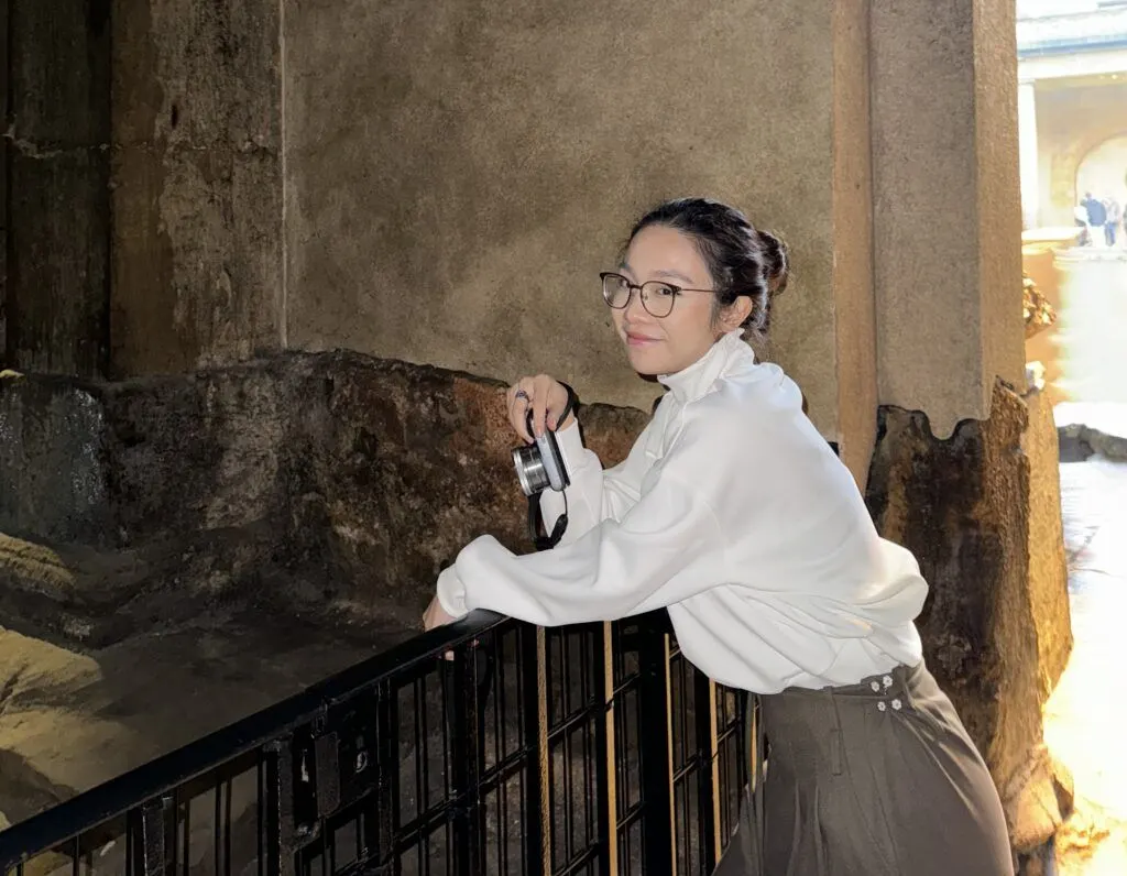 A woman in a white blouse and glasses stands on a staircase, holding a camera and looking to the side.