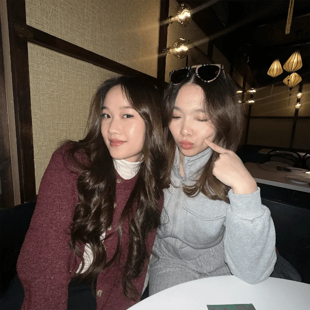 Two young women pose together indoors at night, smiling and making a relaxed gesture near a table.