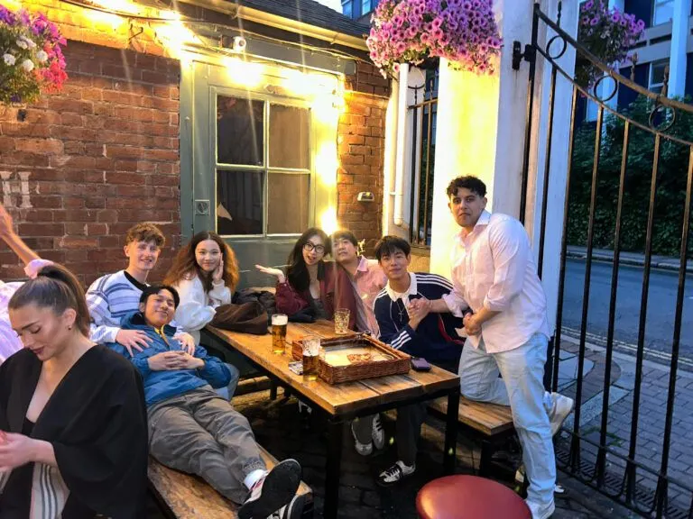 A group of students gathered around an outdoor wooden table at night, with one person standing and speaking while others sit and listen.