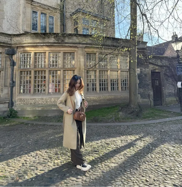 A Vietnamese female student stands outdoors on a cobblestone path in front of an old European-style building, wearing a long coat and carrying a bag.