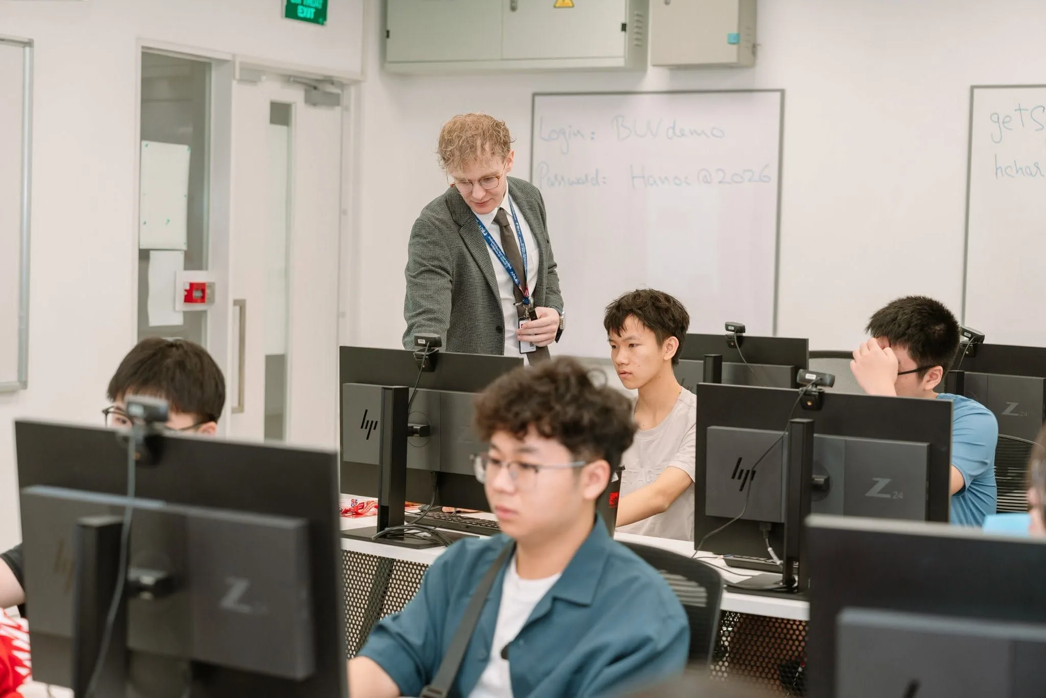A lecturer guiding a student working at a desktop computer in a classroom.