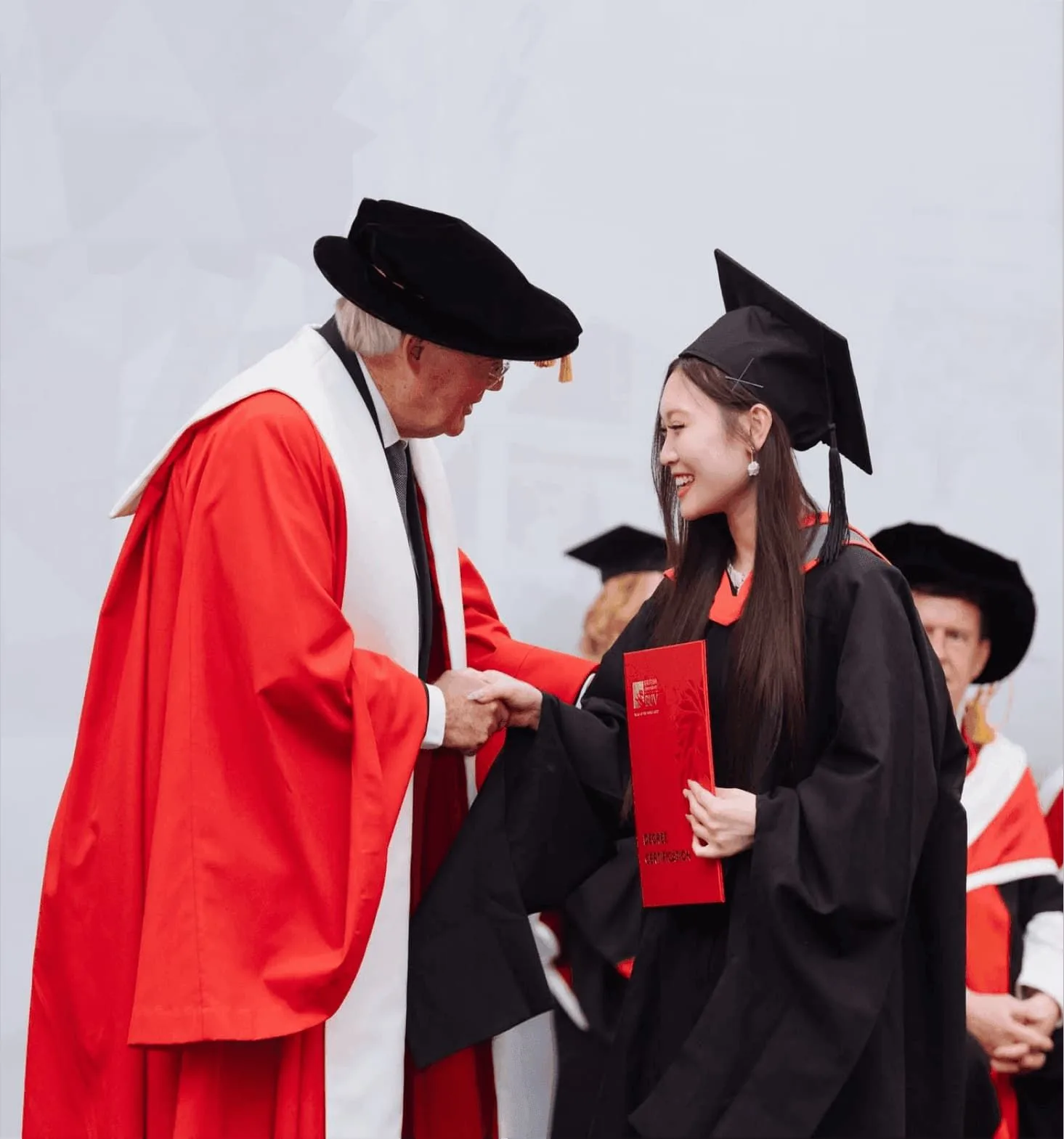 A female graduate in a black cap and gown shakes hands with an academic official in red robes while holding a red diploma folder at a graduation ceremony.
