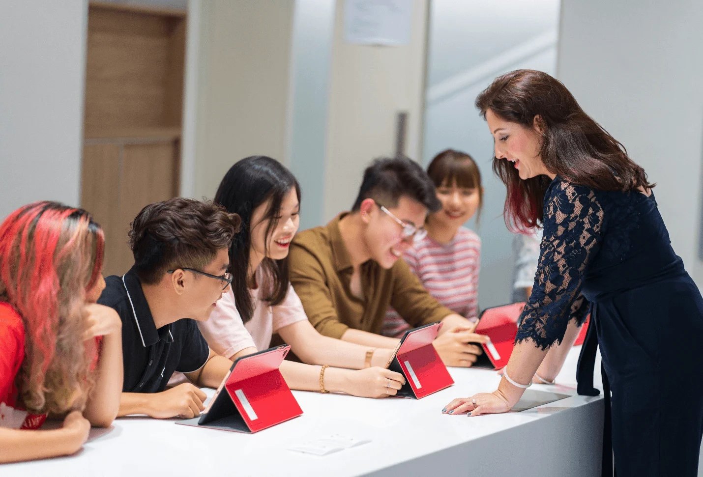 A lecturer engages with five university students gathered around a table, using red tablets during a collaborative classroom activity.