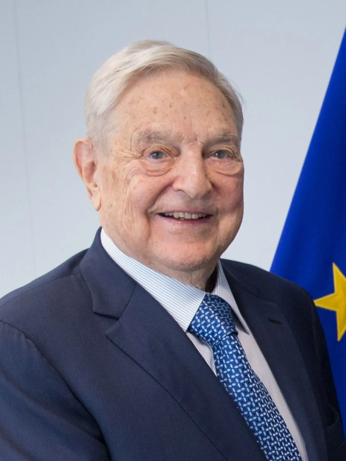 Portrait of George Soros wearing a dark suit and patterned blue tie, with a blue flag featuring yellow stars in the background.