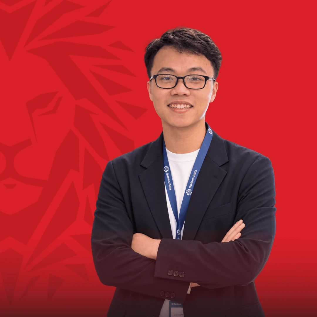 Portrait of a smiling BUV alumnus wearing glasses, a dark blazer, and a blue lanyard, standing with arms crossed against a red background.